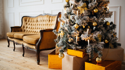 A bright elegant Christmas tree next to a mustard sofa in a bright interior. Christmas gift boxes under the tree. Christmas