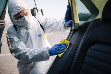 Sanitizing service worker cleans the car's interior with a yellow rug. A man in a protective suit, mask and gloves disinfects the vehicle's doors. Coronavirus covid spread prevention concept.