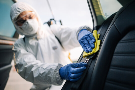 Sanitizing service worker cleans the car's interior with a yellow rug. A man in a protective suit, mask and gloves disinfects the vehicle's doors. Coronavirus covid spread prevention concept.
