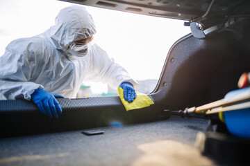 Sanitiry worker disinfects the car's trunk with a yellow rug. A man wearing protective suit, mask...