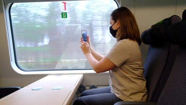 A woman in a black protective mask is sitting alone on the train and taking a picture with the smartphone.