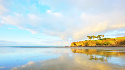 Orange cliff with tall trees on the coast of bay of Douarnenez. Blue sky with colorful evening clouds. Reflections on the water. Brittany, France