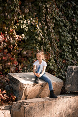 Fototapeta premium Street photo of a little beautiful girl in jeans and a white t-shirt on a background of concrete slabs and autumn leaves