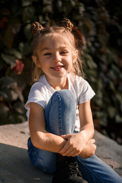 Street photo of a little beautiful girl in jeans and a white t-shirt on a background of concrete slabs and autumn leaves