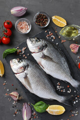 Two raw fish dorado with ingredients and seasonings like basil, lemon, salt, pepper, cherry tomatoes and olive oil on dark background, top view