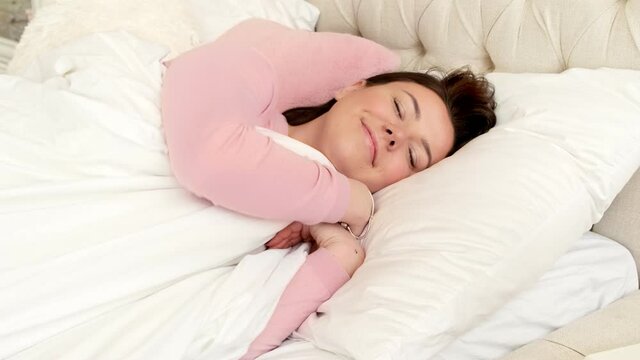 Young woman falling asleep smiling in comfortable bed with fresh linens on a comfortable orthopedic pillow and mattress. Healthy good sleep nap concept. Sleeps in bed in the morning on a weekend.