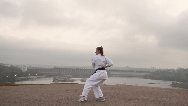 The girl warms up, does karate yoga against the background of a bridge, river, dam, hydroelectric power station. In the fall, cloudy dawn. Overall plan. Static camera