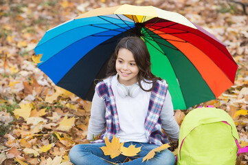 perfect autumn day of cheerful child under colorful umbrella with school bag sitting in fall season...