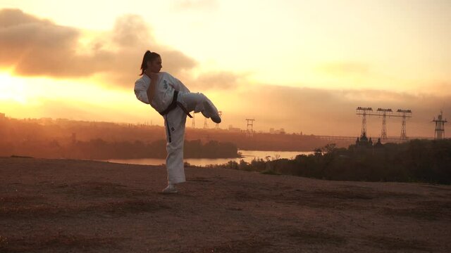 The girl warms up, trains stretching, kick in karate. Harmony with nature. Autumn at sunset. In the background, the golden river, the sun forest, the bridge, the city, the church, the clouds