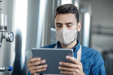 Fototapeta na wymiar Young attractive male worker in protective mask looks at digital tablet and stands near large metal tank