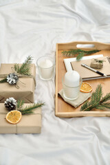 Cosy home flat lay composition with present boxes, blank copybook, a pen, tea and cones on a bed. Holiday shopping list, wish list or New Year Goals concept, presents checklist. Selective focus