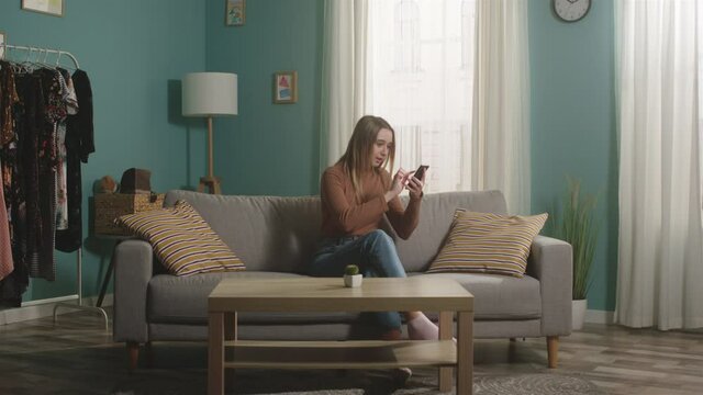 Young girl in blue jeans and a brown sweater sits on a beige sofa, looks at something on the phone and openly expresses joyful emotions