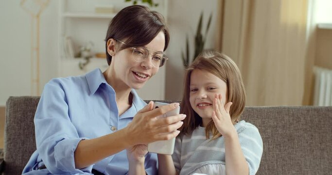 Young mother with glasses sister nanny sitting with her daughter girl looking at phone together using smartphone make photo or video. Child lady eating apple hugs mom looks at screen of mobile device