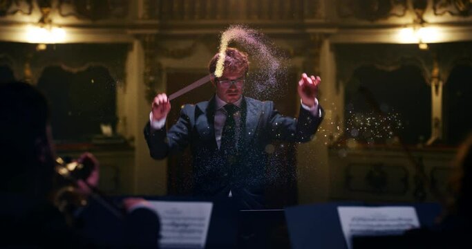 Cinematic close up shot of conductor directing with magic glows symphony orchestra with performers playing violins,cello and trumpet on classic theatre stage during music concert with dramatic lights.