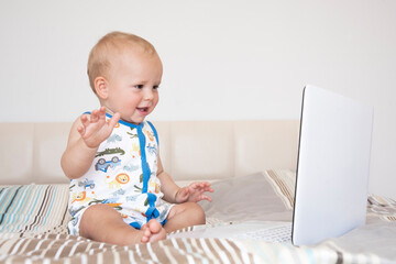 Funny cute child boy sits on a bed in front of an open computer laptop and emotionally looks at the screen.