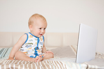 Adorable cute child boy sits on a bed in front of an open computer laptop and emotionally looks at the screen.

