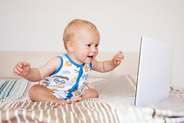 Adorable cute child boy sits on a bed in front of an open computer laptop and emotionally looks at the screen.

