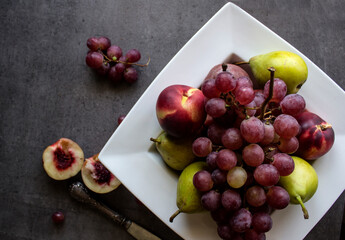 Juicy fruits on white plate.  Top view photo of fresh nectarines, pears and grapes. Gray textured background. Fruits diet. 