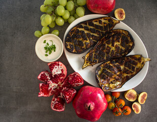 Roasted eggplants on white plate, pomegranates and grapes on a table. Mediterranean style food. 