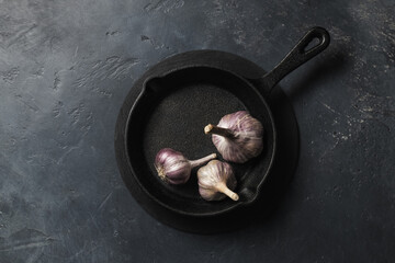Small cast iron frying pan with farm garlic on dark concrete background, place for text