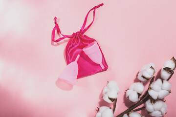 Two pink menstrual cups on a pink cloth bag and a branch of cotton on a pink background
