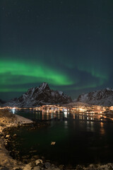 Fototapeta na wymiar Dancing green polar lights over the village Reine on the Lofoten islands in Norway at night in winter with snow capped mountains