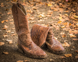 A pair of brown cowboy boots,one fallen over, found on a leaf covered path.