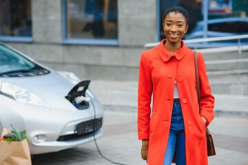 Young african woman is standing near the electric car and looking at camera. The rental car is...