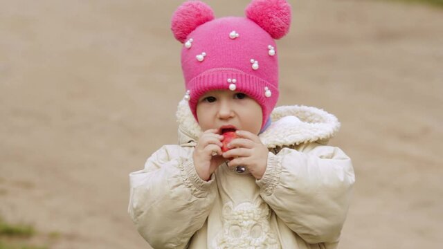 A two-year-old girl in a raspberry cap is eating an Apple on the street.