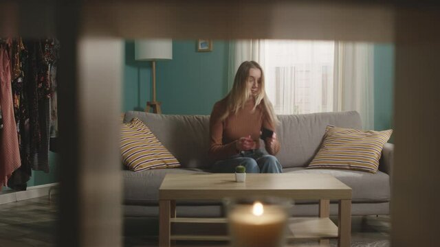 Young lady in blue jeans and brown sweater walks into room, sits down on sofa and picks up phone. Directions camera right. In foreground mezzanine with lighted candle.