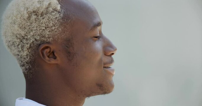 Side view african man afro american guy teenager blond student standing outdoors with eyes closed deeply inhaling fresh warm cold air with his nose enjoying calm life summer weather, smiling toothy