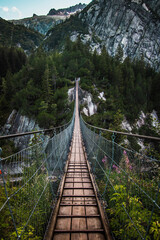 Wooden long footbridge above the deep gorge with a river beach at the bottom, between the rocks. Wild mountains in Swiss Alps