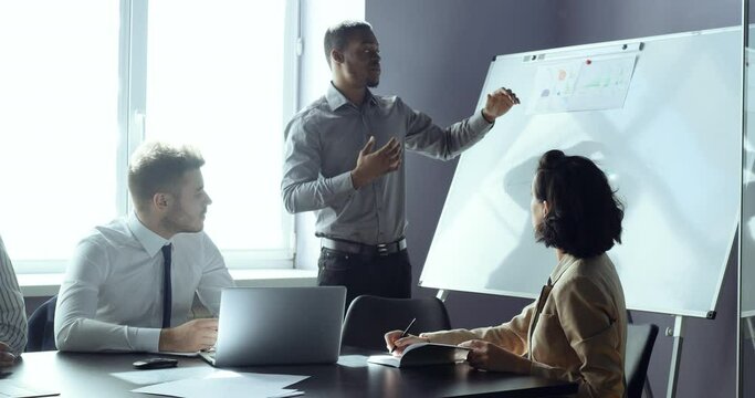 African business man team leader presenting project strategy showing ideas on whiteboard in office for colleagues, three people coworkers sitting at table in conference enjoying training seminar