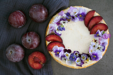 Cheesecake decorated with red plums and edible flowers. Dark background. Top view photo of homemade dessert. 
