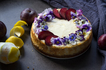 Cheesecake decorated with red plums and edible flowers. Fabric texture background. Top view photo of homemade dessert. 
