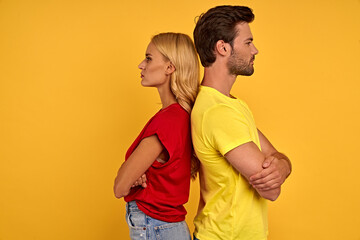 Side view of smiling young couple in red and yellow t-shirts isolated on yellow background. People lifestyle concept. Mock up copy space. Standing back to back holding hands crossed.