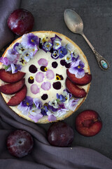 Cheesecake decorated with red plums and edible flowers. Dark background. Top view photo of homemade dessert. 
