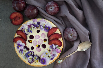 Cheesecake decorated with red plums and edible flowers. Dark background. Top view photo of homemade dessert. 
