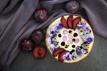 Cheesecake decorated with red plums and edible flowers. Dark background. Top view photo of homemade dessert. 
