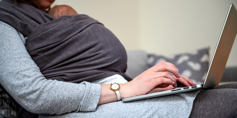 Woman working from home on laptop with her infant baby in the sling during Covid-19 pandemic - modern teleworking