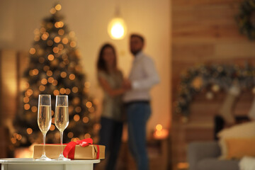 Glasses with champagne and Christmas gift on table and couple in room, bokeh effect. Space for text