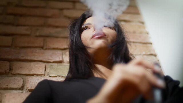 Dark-haired young woman smoking electronic cigarette on brick wall background
