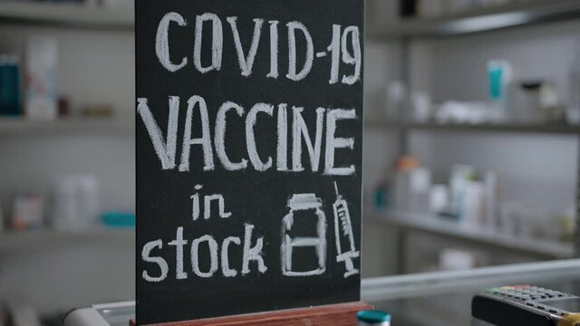 A chalk plaque with the inscription about the presence of a vaccine for coronavirus covid-19 in the interior of the pharmacy. A ampule with a vaccine stands on the window in front of the sign.