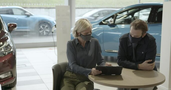 Saleswoman advising male on car purchase in vehicle dealership showroom using digital tablet
