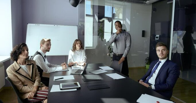 Multiethnic group of partners sitting in corporate meeting room, male african analyst uses digital interactive whiteboard for presentation of project, lawyers investors listen report looking at screen