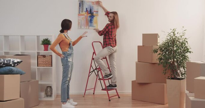 Handsome young guy standing on ladder holding picture. Beautiful Caucasian girl gives advice on where to hang new curtain in apartment.