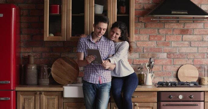 Married couple young wife sit on countertop hug husband family using tablet device search kitchen interior design ideas on internet, choose remodelling services online spend free time together at home