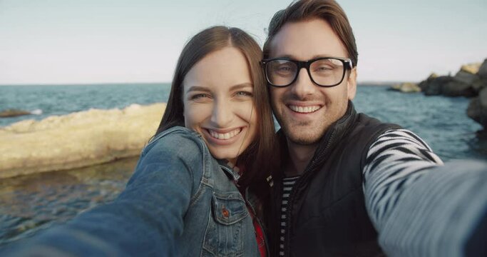 Cheerful young couple holding camera and making grimaces while having fun. Two happy people spend time together by sea. Camera view.