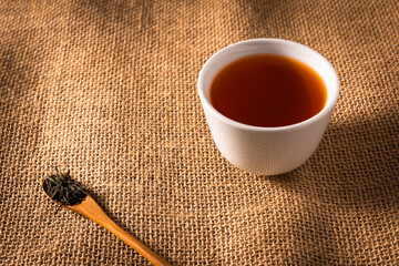 A cup of hot Chinese Oolong tea in a white tea cup put on sackcloth with a wooden teaspoon full of...