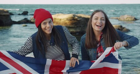 Two young happy female countrymen walking along the beach holding flag of Great Britain. Exchange student. Travelers. - Powered by Adobe